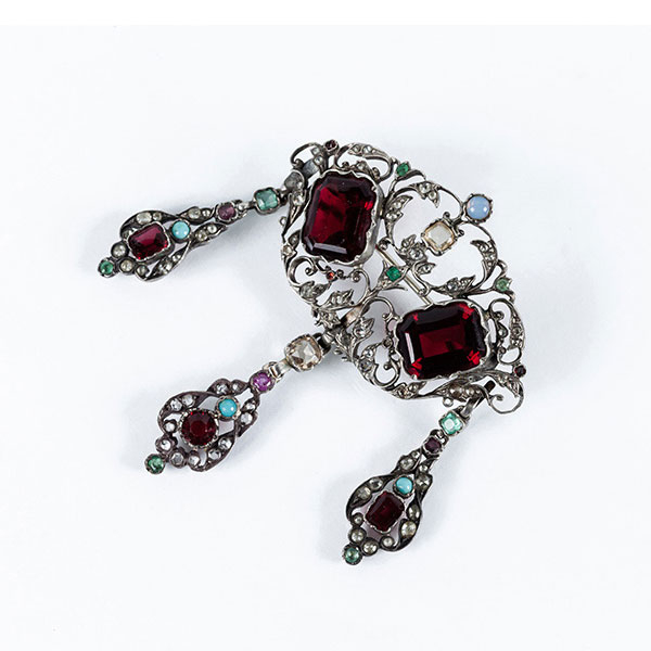 Alfiler antiguo (s XIX) en montura de plata con dos grandes piedras de color granate sobre celosía calada con motivos vegetales decorados con diamantitos, esmeralditas y piedritas de color.