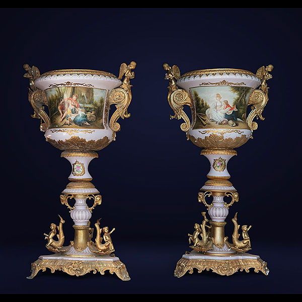 Porcelana y bronce dorado. Dimensiones: 125 x 61 x 59 cm Conjunto decorativo de dos grandes jarrones de porcelana fina decorados con escenas mitológicas y ornamentados con detalles en bronce dorado. Cada jarrón está ricamente adornado con relieves dorados y asas en forma de querubines. La policromía detallada y el diseño elegante reflejan la influencia de la manufactura Sèvres y el gusto neoclásico francés.