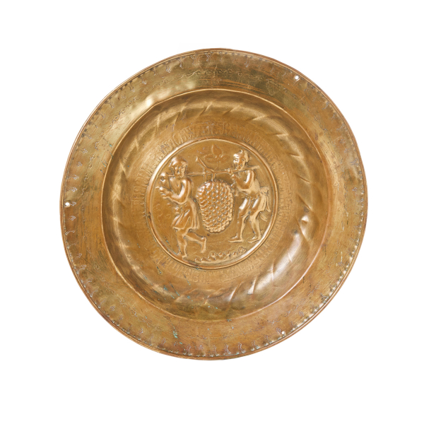 Plato petitorio de limosna en latón repujado con decoración central de escena con los espías de Canaan, Josua y Caleb portando las uvas custodiada por cenefa con letras góticas, Núremberg, Alemania, s.XV.