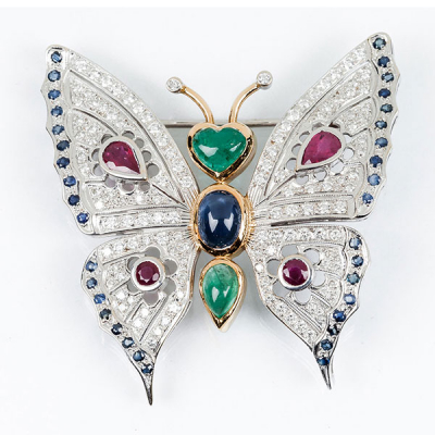 Bello alfiler en montura calada de oro blanco y amarillo 18 K, en forma de mariposa con esmeraldas, zafiros, rubíes y alas cuajadas de limpios y blancos diamantes, talla brillante 